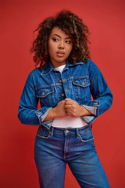 Fashionable young african american woman in stylish denim outfit looking away on red backdrop — Stock Photo