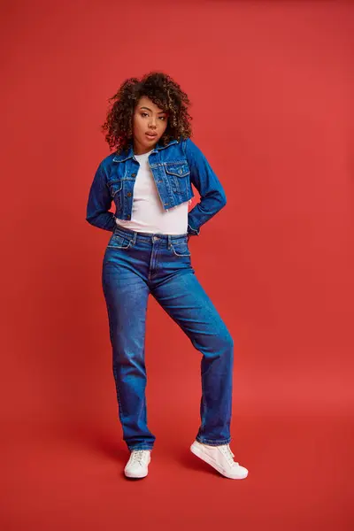 Приваблива шикарна афроамериканська жінка в джинсовому одязі позує на червоному тлі і дивиться на камеру — стокове фото