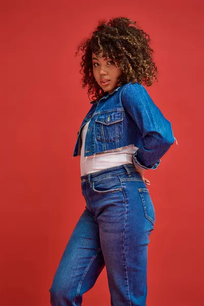 Витончена афроамериканська жінка в джинсовому одязі позує на червоному тлі і дивиться на камеру — стокове фото