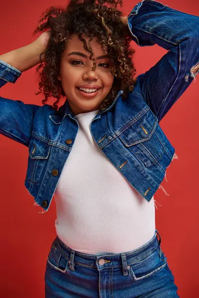 Cheerful african american woman in denim attire posing on red backdrop and looking at camera — Stock Photo