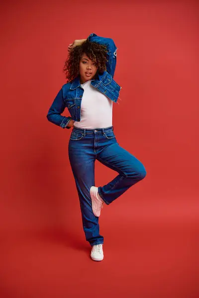 Good looking african american woman in denim attire posing on red backdrop and looking at camera — Stock Photo