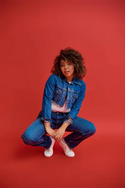 Stylish african american fashionista in denim attire posing on red backdrop and looking at camera — Stock Photo