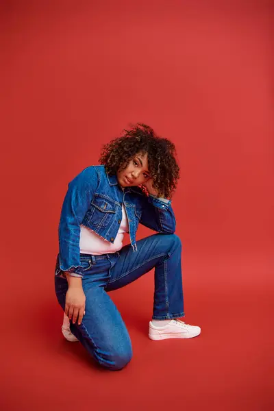 Trendy african american fashionista in denim attire posing on red backdrop and looking at camera — Stock Photo