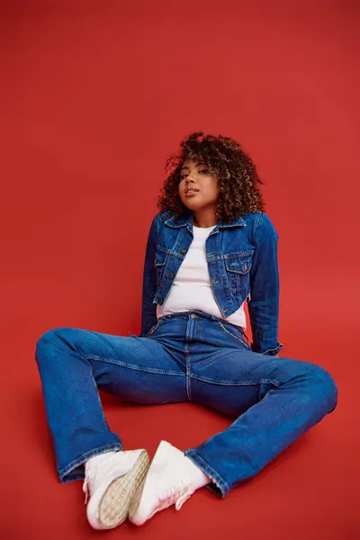 Elegant african american fashionista in denim attire posing on red backdrop and looking at camera — Stock Photo