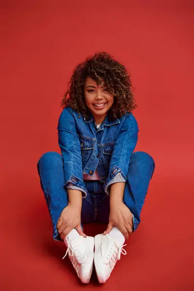 Joyful african american female model in denim attire looking at camera and posing on red background — Stock Photo
