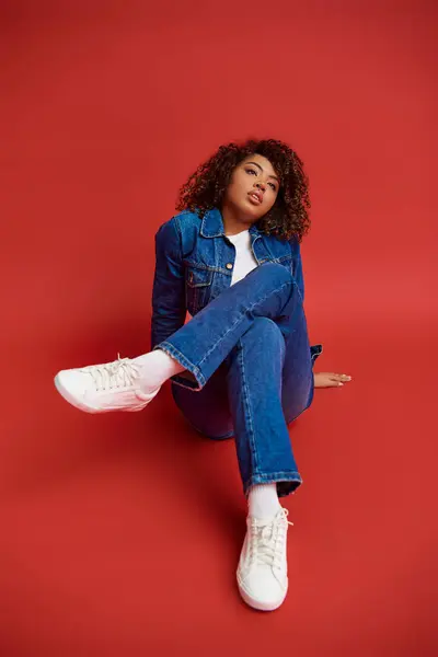 Alluring young african american woman in stylish denim outfit looking away on red backdrop — Stock Photo