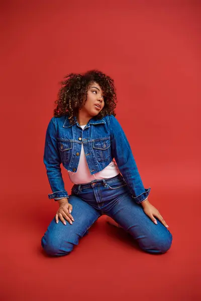 Attractive young african american woman in stylish denim outfit looking away on red backdrop — Stock Photo