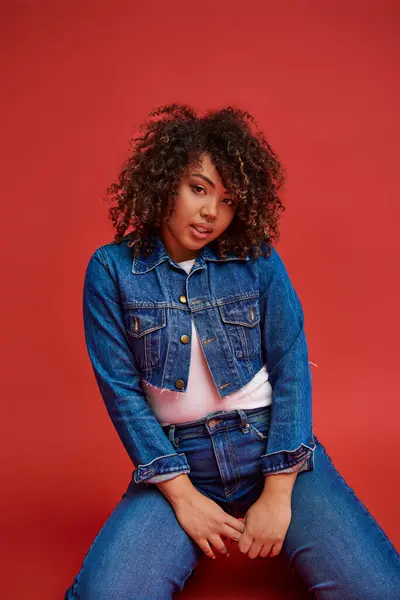 Exquisite african american female model in denim attire looking at camera and posing on red backdrop — Stock Photo