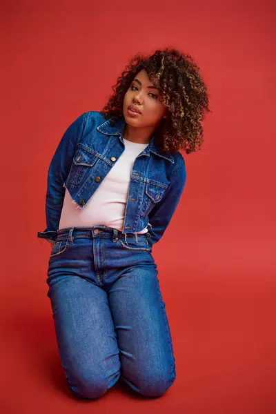 Refined african american female model in denim attire looking at camera and posing on red background — Stock Photo