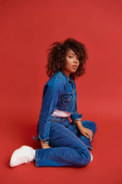 Refined african american woman in stylish denim outfit looking at camera on vibrant red backdrop — Stock Photo