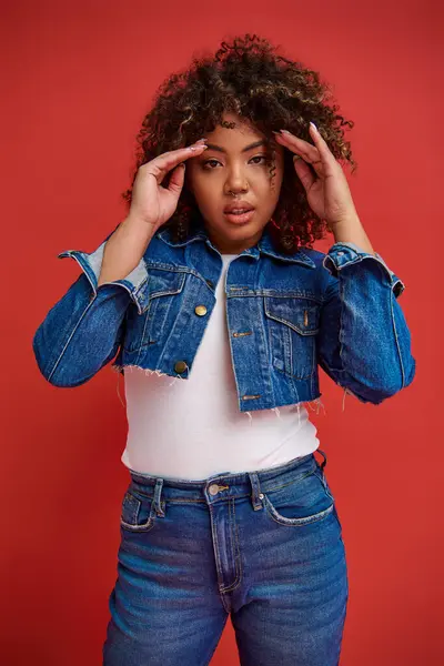 Graceful african american woman in stylish denim outfit looking at camera on vibrant red backdrop — Stock Photo