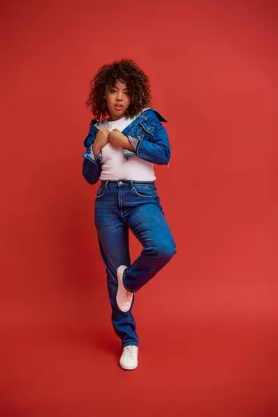 Елегантна афроамериканка в стильному джинсовому вбранні дивиться на камеру на яскравому червоному тлі — стокове фото