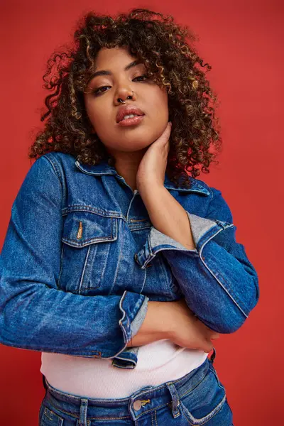 Attractive african american woman in stylish denim outfit looking at camera on vibrant red backdrop — Stock Photo