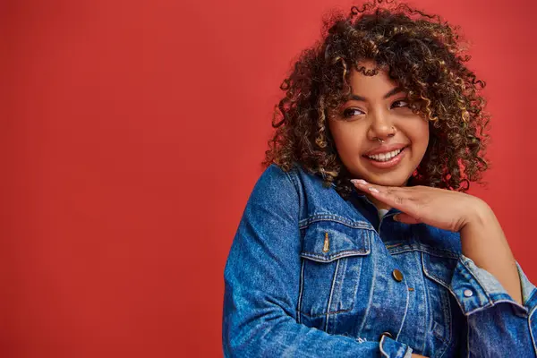 Joyous appealing african american woman in stylish denim outfit looking away on red backdrop — Stock Photo