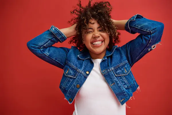 Веселая африканская американка в джинсовой одежде счастливо позирует с закрытыми глазами на красном фоне — стоковое фото