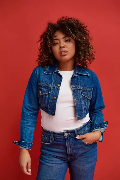Alluring african american woman in stylish denim outfit looking at camera on vibrant red backdrop — Stock Photo