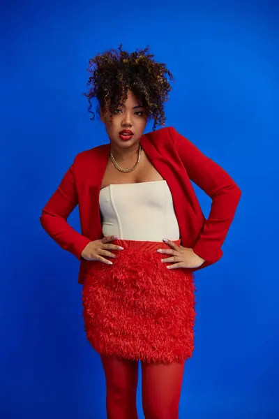 Mujer afroamericana de moda en falda roja y chaqueta posando sobre fondo azul y mirando a la cámara - foto de stock
