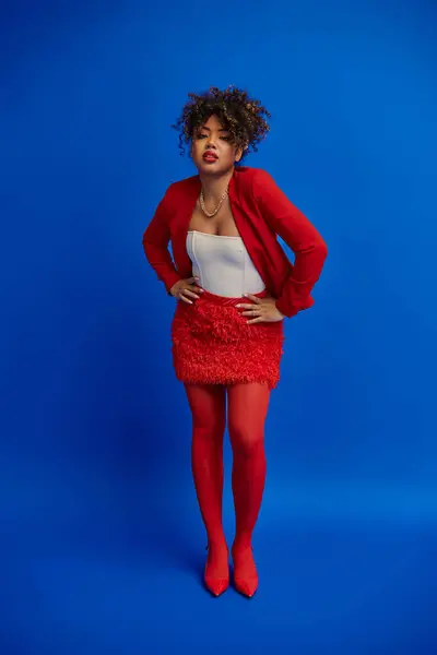 Femme afro-américaine élégante en jupe rouge et blazer posant sur fond bleu et regardant la caméra — Photo de stock