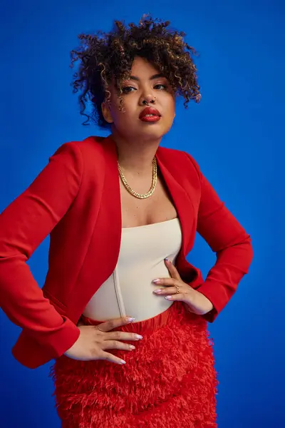 Graceful african american woman in red skirt and blazer on blue backdrop and looking at camera — Stock Photo