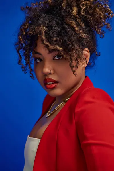 Young african american woman in red skirt and blazer posing on blue backdrop and looking at camera — Stock Photo