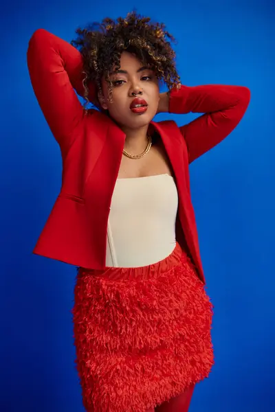 Elegante junge Afroamerikanerin im roten Blazer mit lockigem Haar blickt in die Kamera, blauer Hintergrund — Stockfoto