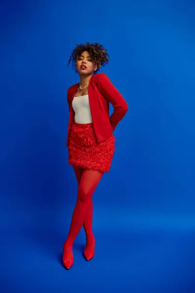 Trendy young african american woman in red blazer with curly hair looking at camera, blue backdrop — Stock Photo