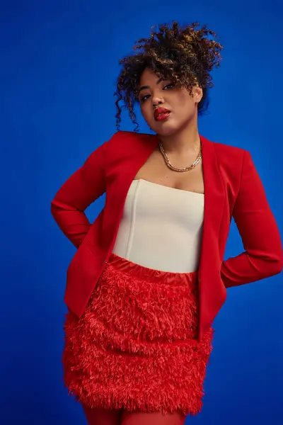 Dapper young african american woman in red blazer with curly hair looking at camera, blue backdrop — Stock Photo