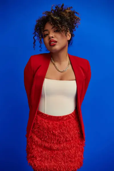 Exquisita mujer afroamericana elegante en chaqueta roja con pelo rizado mirando a la cámara, telón de fondo azul - foto de stock