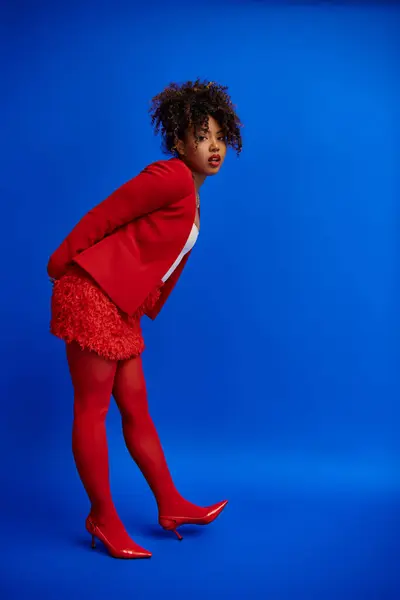 Tasteful chic african american woman in red blazer with curly hair looking at camera, blue backdrop — Stock Photo