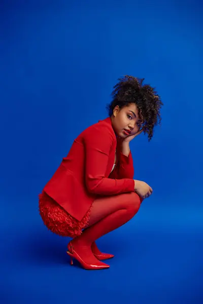 Polished chic african american woman in red blazer with curly hair looking at camera, blue backdrop — Stock Photo