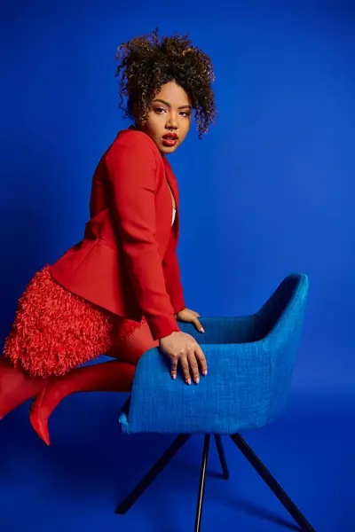 Sexy elegante mulher americana africana em terno vermelho inteligente sentado na cadeira azul e olhando para a câmera — Fotografia de Stock