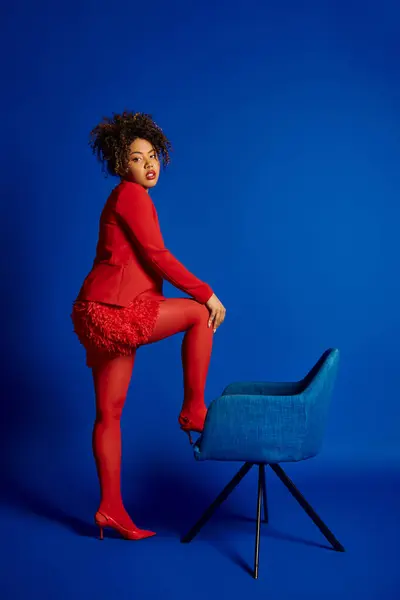 Приваблива шикарна афроамериканська жінка в смарт-червоному костюмі, сидячи на синьому стільці і дивлячись на камеру — стокове фото