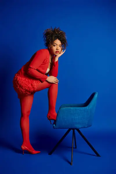 Bella donna africana americana chic in abito rosso intelligente seduto sulla sedia blu e guardando la fotocamera — Foto stock