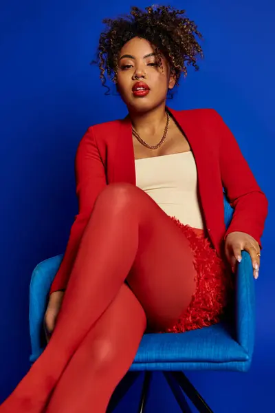Encantadora mulher americana africana chique em terno vermelho inteligente sentado na cadeira azul e olhando para a câmera — Fotografia de Stock