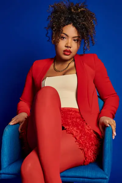 Charmante afrikanische Amerikanerin im schicken roten Anzug sitzt auf blauem Stuhl und blickt in die Kamera — Stockfoto