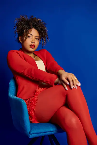Гламурна шикарна афроамериканська жінка в смарт-червоному костюмі, сидячи на синьому стільці і дивлячись на камеру — стокове фото
