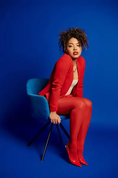 Enticing chic african american woman in smart red suit sitting on blue chair and looking at camera — Stock Photo