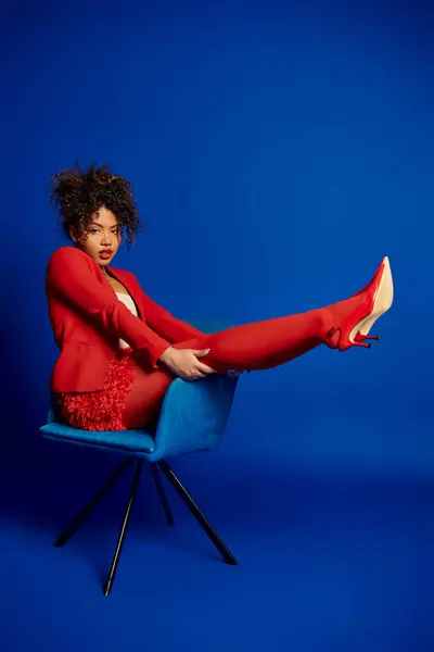 Verführerisch schicke afrikanisch-amerikanische Frau im schicken roten Anzug sitzt auf blauem Stuhl und blickt in die Kamera — Stockfoto