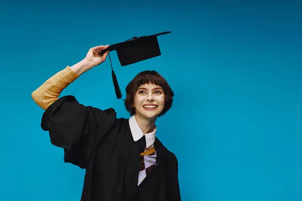 Happy college girl wearing black graduation gown holding academic cap on blue background in studio — Stock Photo