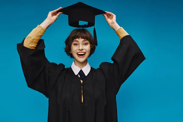 Sonriente chica universitaria con vestido de graduación negro celebración de la tapa académica sobre fondo azul en el estudio - foto de stock