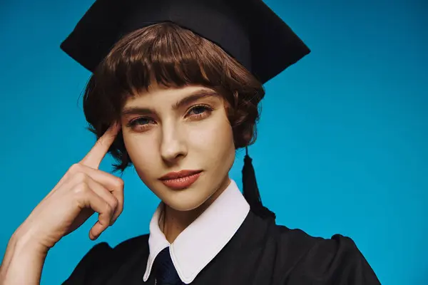 Retrato de chica universitaria pensativa con vestido de graduación negro y gorra académica sobre fondo azul - foto de stock