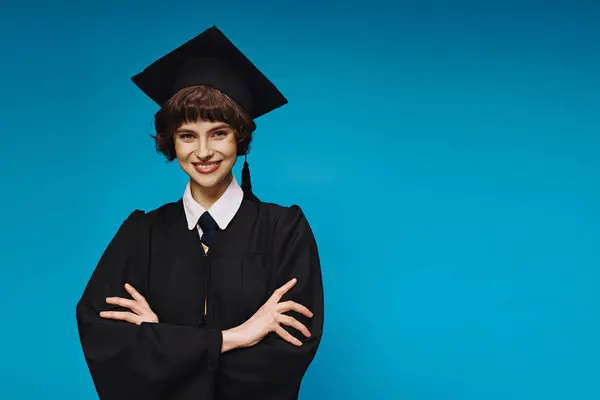 Portrait of smiling college girl in black graduation gown and academic cap posing with crossed arms — Stock Photo
