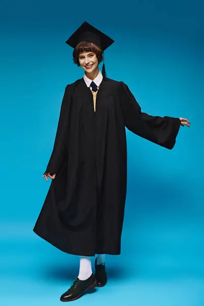 Graduation concept, happy college girl in academic cap and gown standing on blue background — Stock Photo