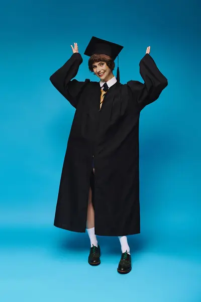 Graduation concept, cheerful college girl in academic cap and gown standing on blue background — Stock Photo