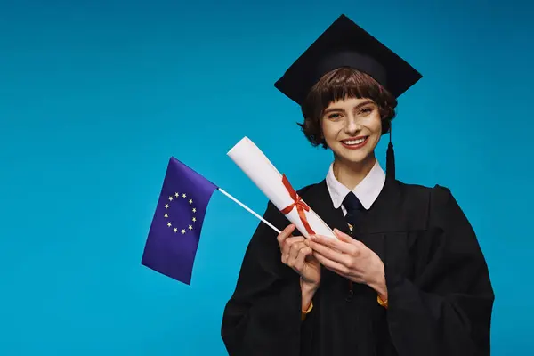 Cheerful graduated college girl in gown and academic cap with diploma and EU flag on blue backdrop — Stock Photo