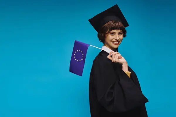 Graduated college girl in gown and academic cap with diploma and EU flag smiling on blue backdrop — Stock Photo