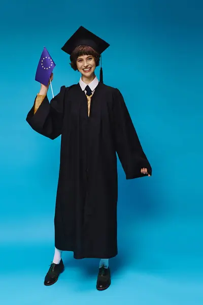 Graduated college girl in gown and academic cap holding EU flag and smiling on blue backdrop — Stock Photo