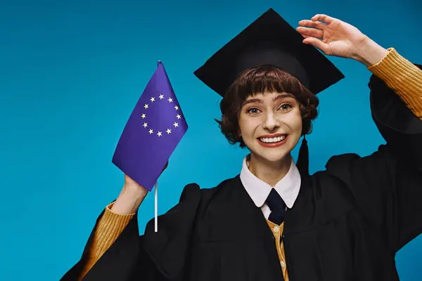 Graduated girl in gown and cap holding EU flag and smiling on blue backdrop, European education — Stock Photo