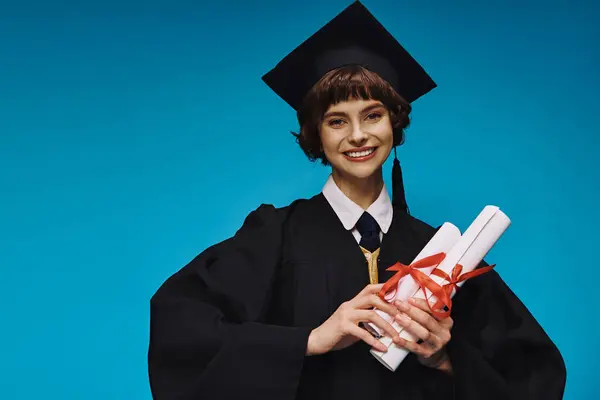 Happy graduated college girl in gown and academic cap holding diplomas with pride on blue — Stock Photo
