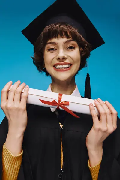 Cheerful grad college girl in gown and academic cap looking at camera with diploma in hands on blue — Stock Photo